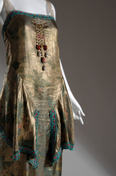 mannequin in a gold lame dress with decorative dangling beads at center bust and draped decorative overskirt with turquoise beaded edge