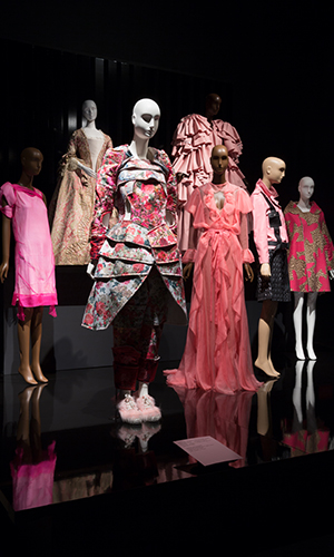 platform view of several pink ensembles including Comme des Garcons armor like tunic with long articulated sleeves in shades of pink and floral jacquard
